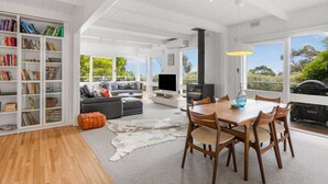 A spacious living area with a wooden dining table, a sofa, and a bookshelf filled with books. - The Deck House Lorne (Lorne)