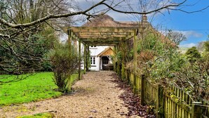 Een landelijke pad dat leidt naar een huis met rieten dak en een houten pergola erboven.