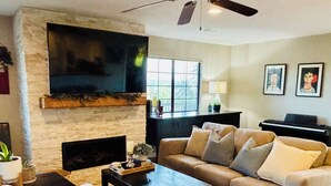 A living room with a fireplace, a ceiling fan, a television mounted on the wall, and a piano. - Perfect 4-bedroom house near lakes and trails in Folsom (Folsom)