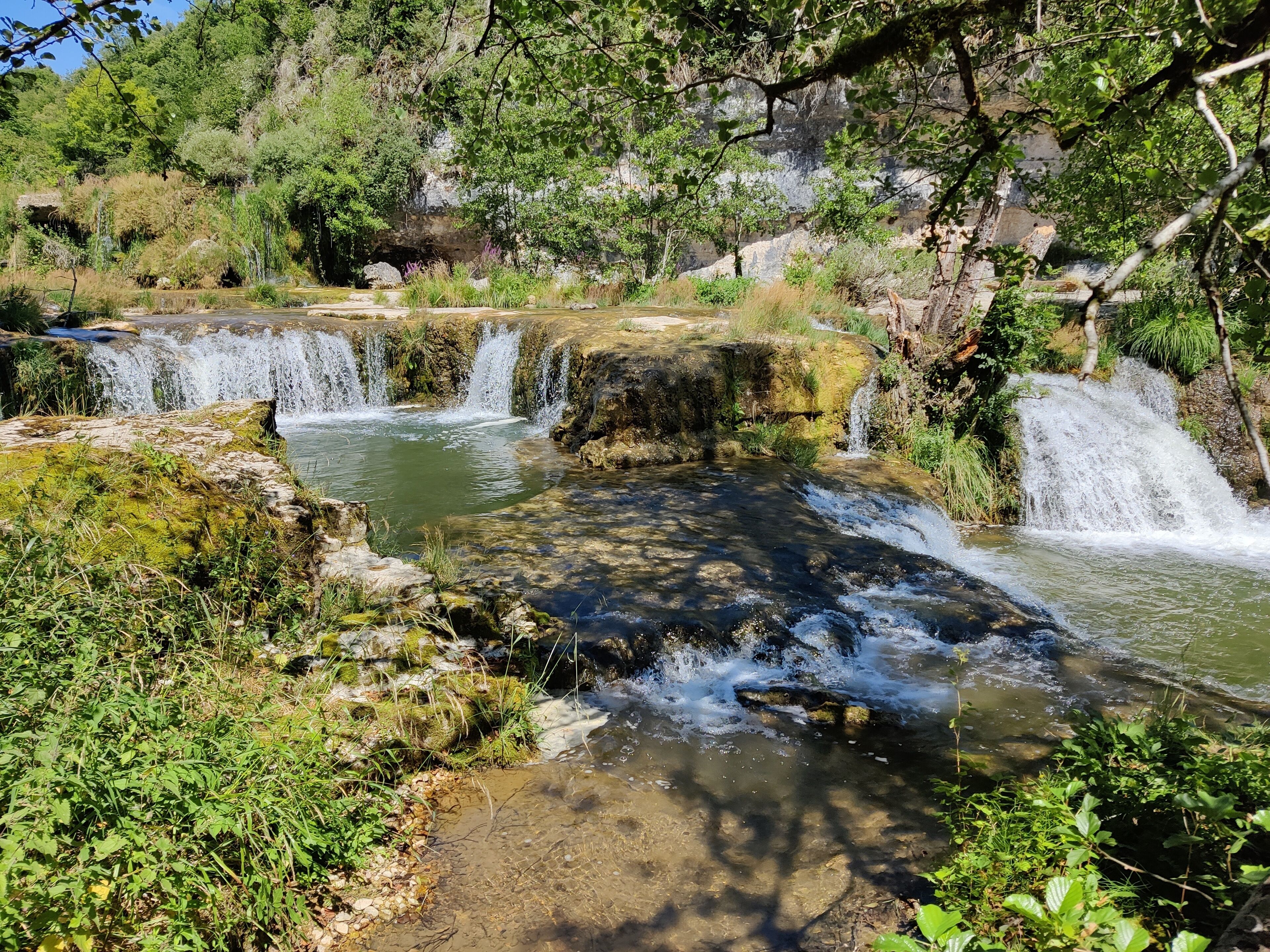 A natural waterfall with clear water flowing over rocks, surrounded by lush greenery and trees.