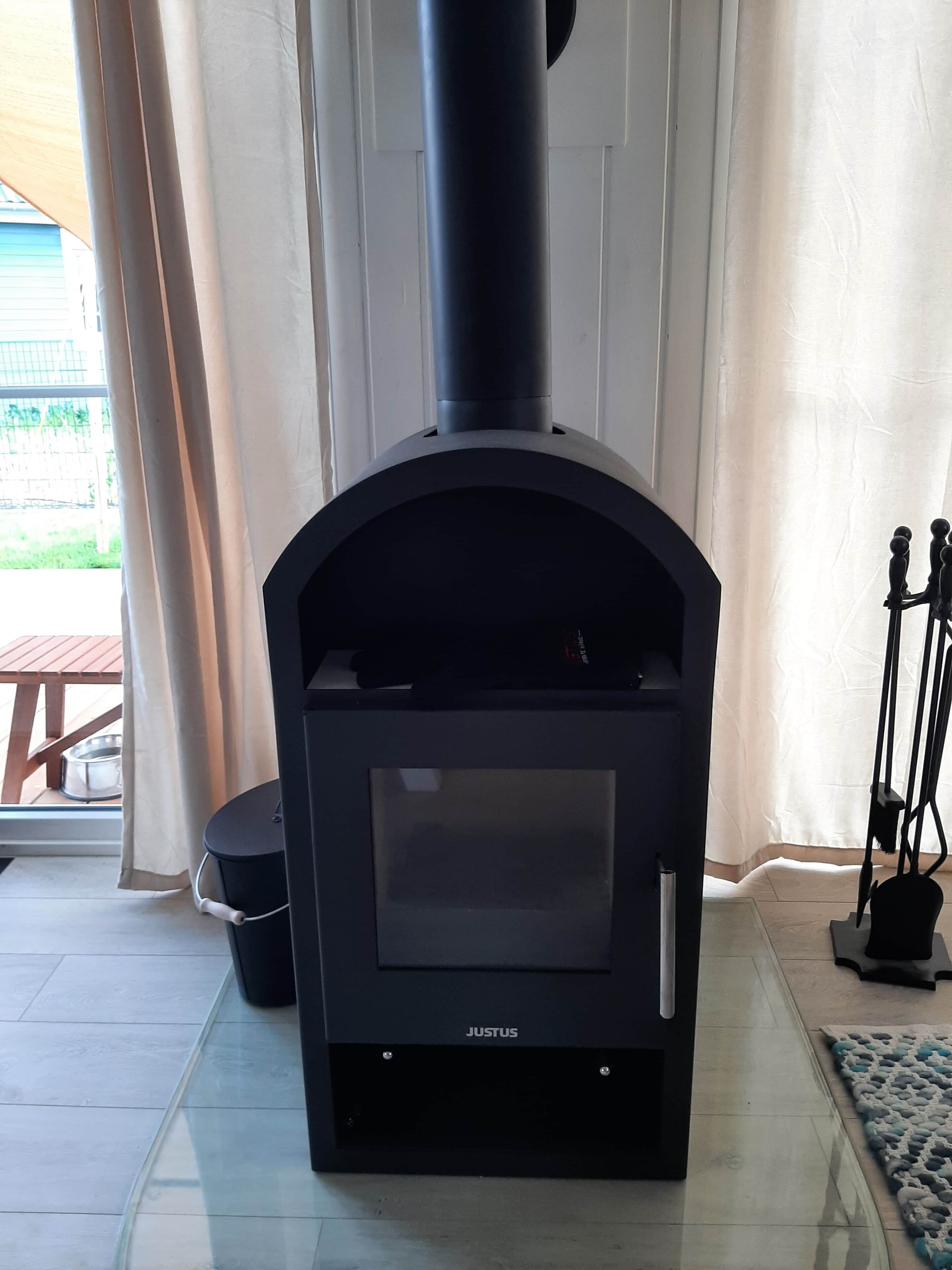 A black wood stove with a glass door, labeled "JUSTUS," in a room with a glass door and a view of a wooden deck outside.