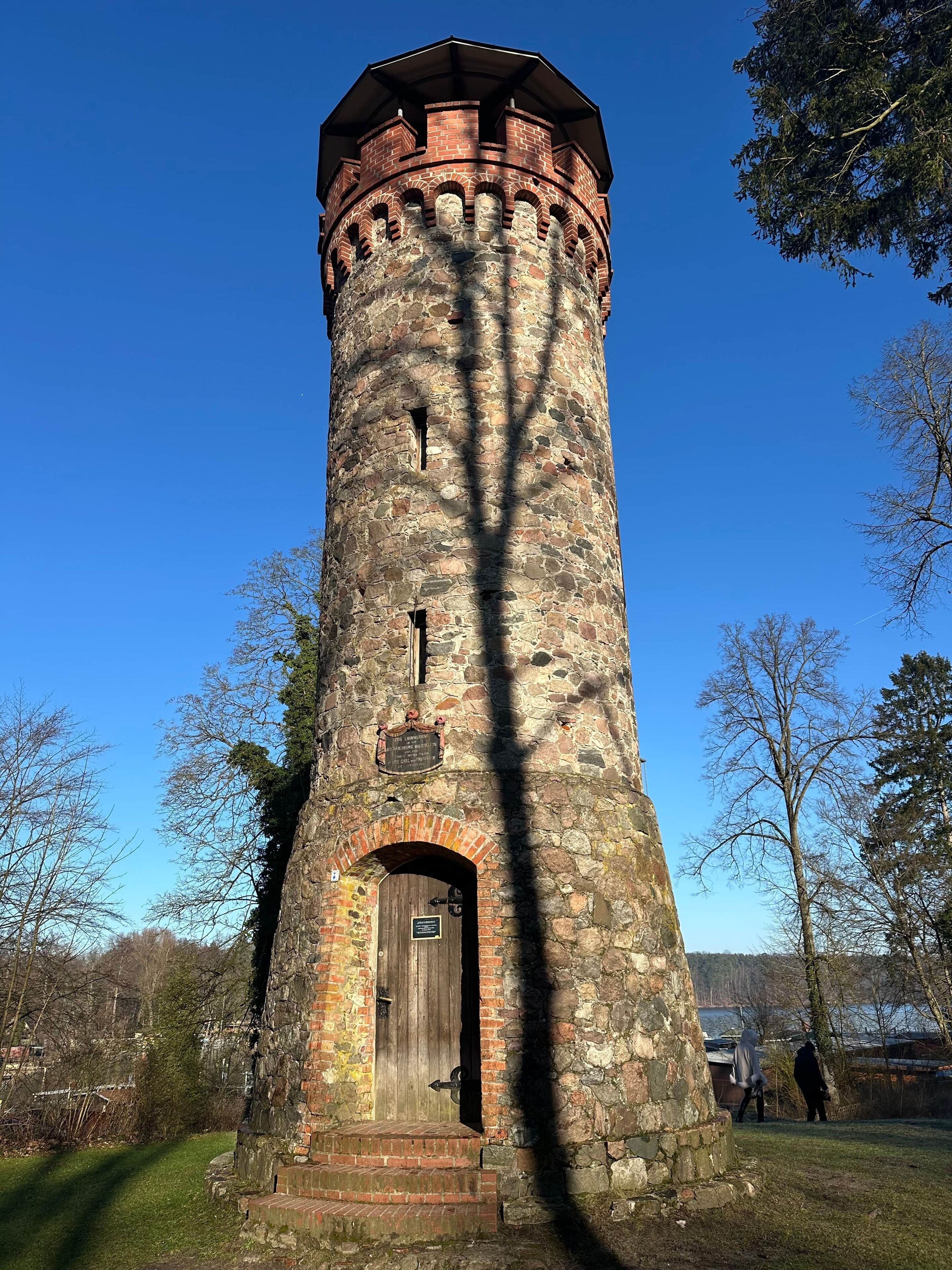 Une tour de pierre avec une porte en bois et une plaque, entourée d’arbres et sous un ciel dégagé.