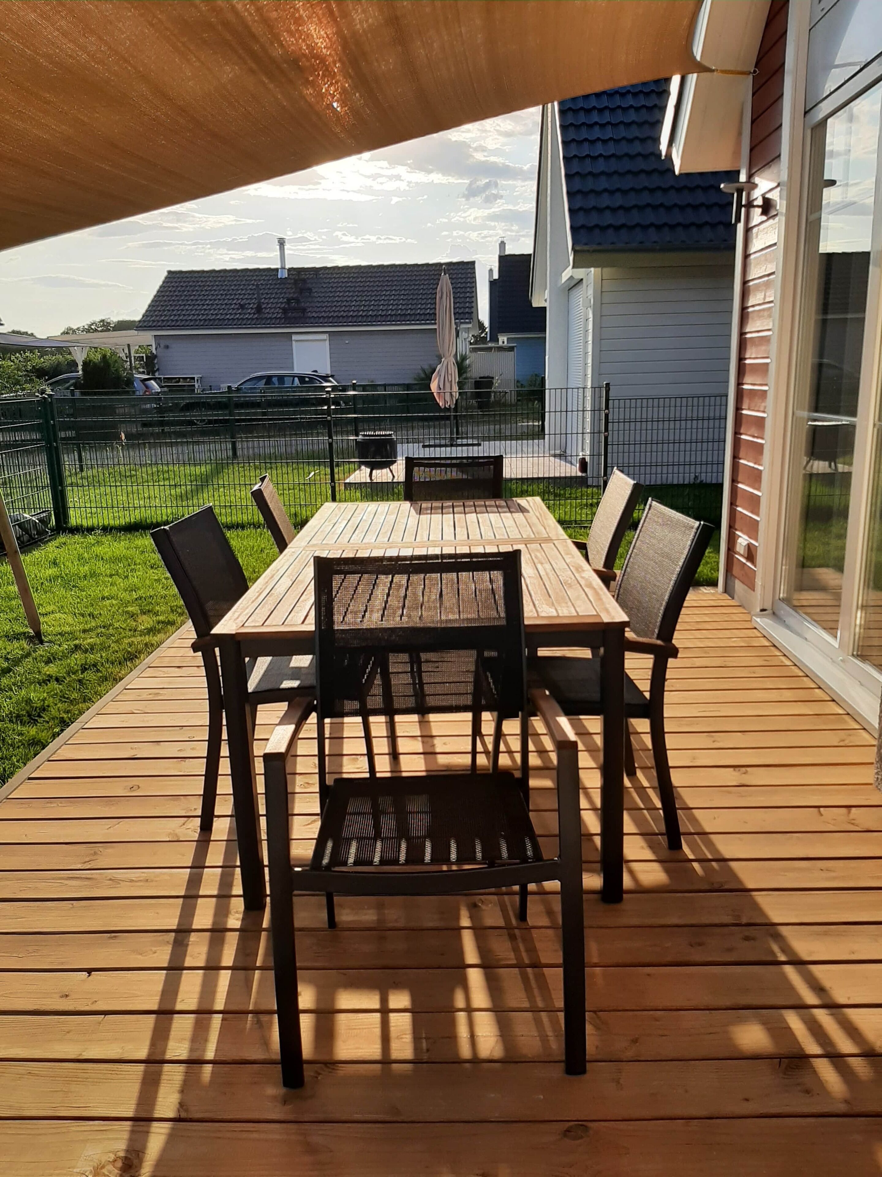 Een houten terras met een tafel en stoelen, een pergola erboven en een uitzicht op een huis en schutting op de achtergrond.