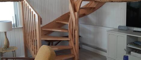 Un escalier en bois, avec un panier tressé suspendu au plafond, un canapé, une table ornée d’un vase de tournesols et un téléviseur posé sur un meuble blanc.