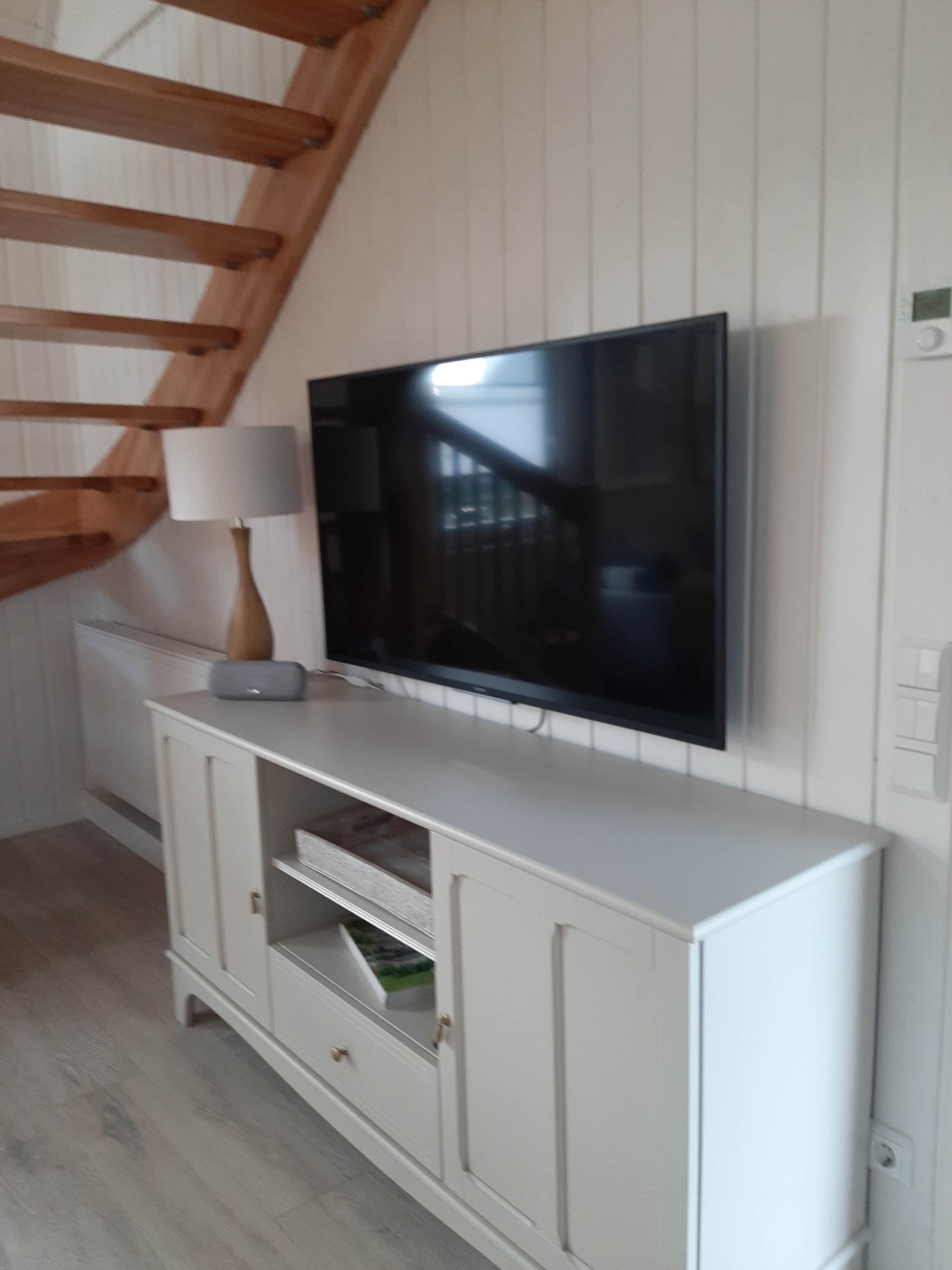 A modern living room with a flat-screen TV mounted on a white wall, a white cabinet below, and a wooden staircase in the background.