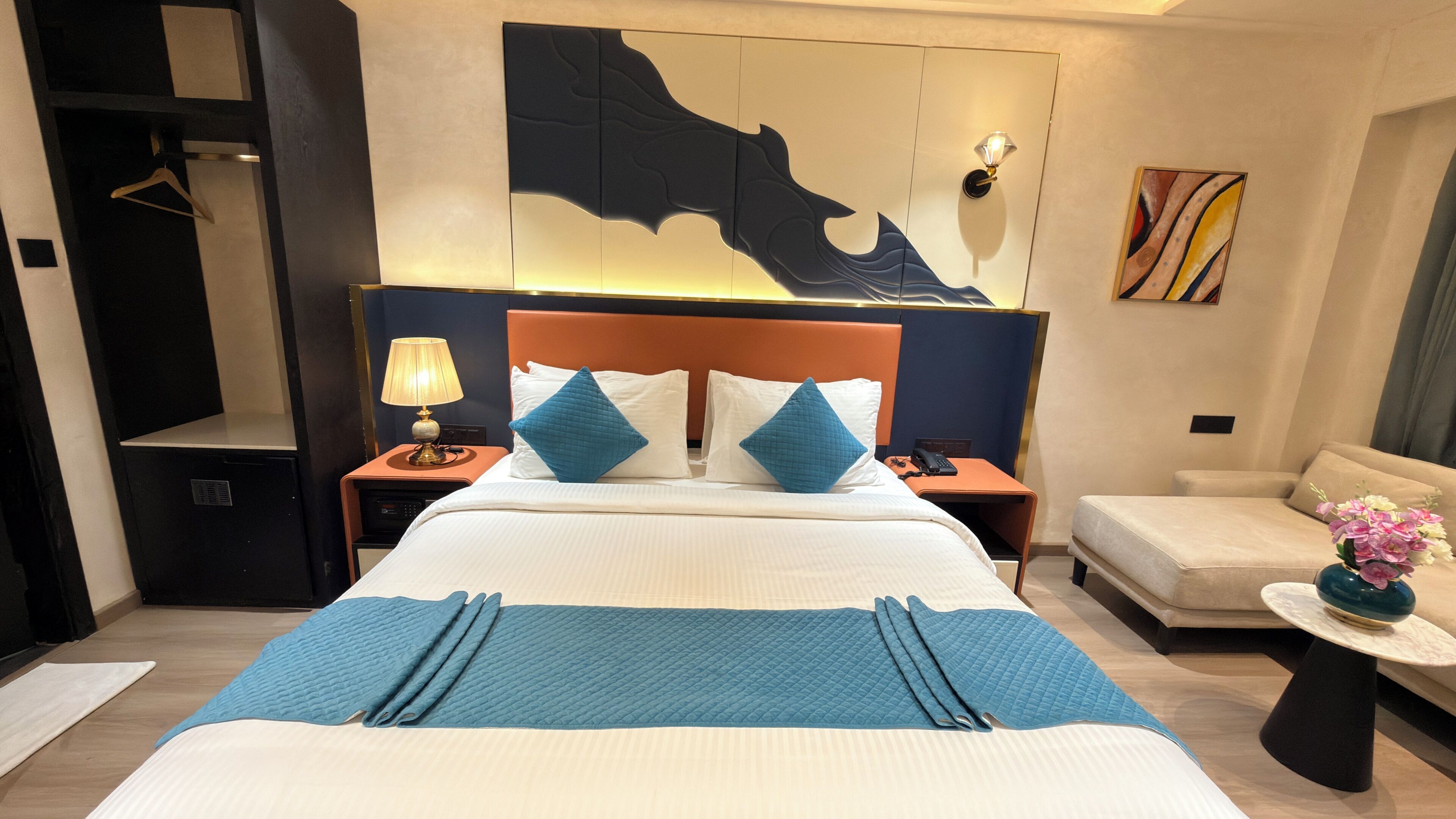 Ein Hotelzimmer mit Bett, Nachttisch mit Lampe, einem Wandbild, einer Couch und einem kleinen Tisch mit Blumen.