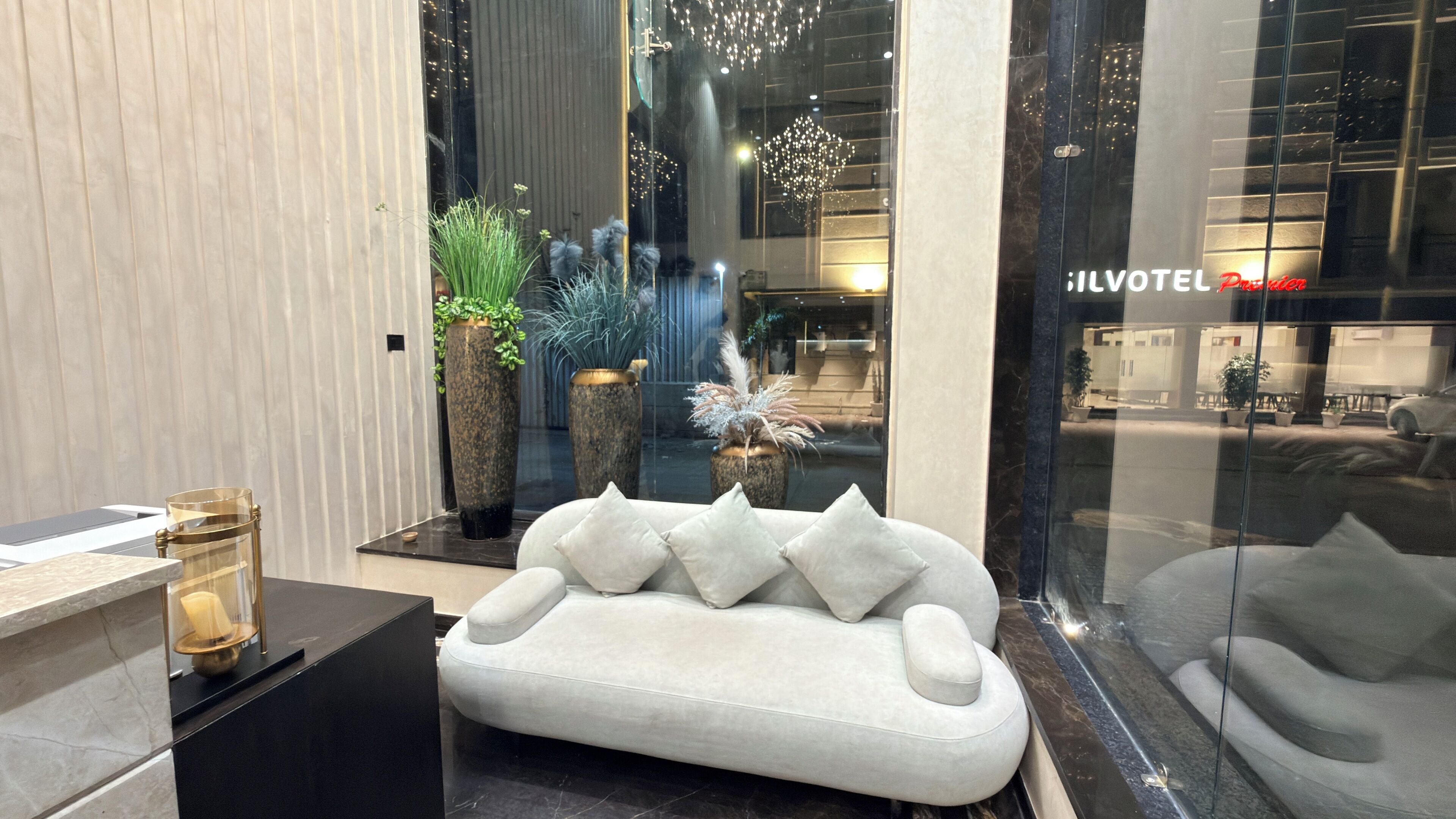 A modern interior with a white sofa, large potted plants, and a glass wall with a reflection of the outside.