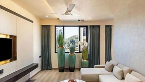 A modern living room with a flat-screen TV, a ceiling fan, a sofa, and large windows with curtains. - Skyearth Hotel (Indore)