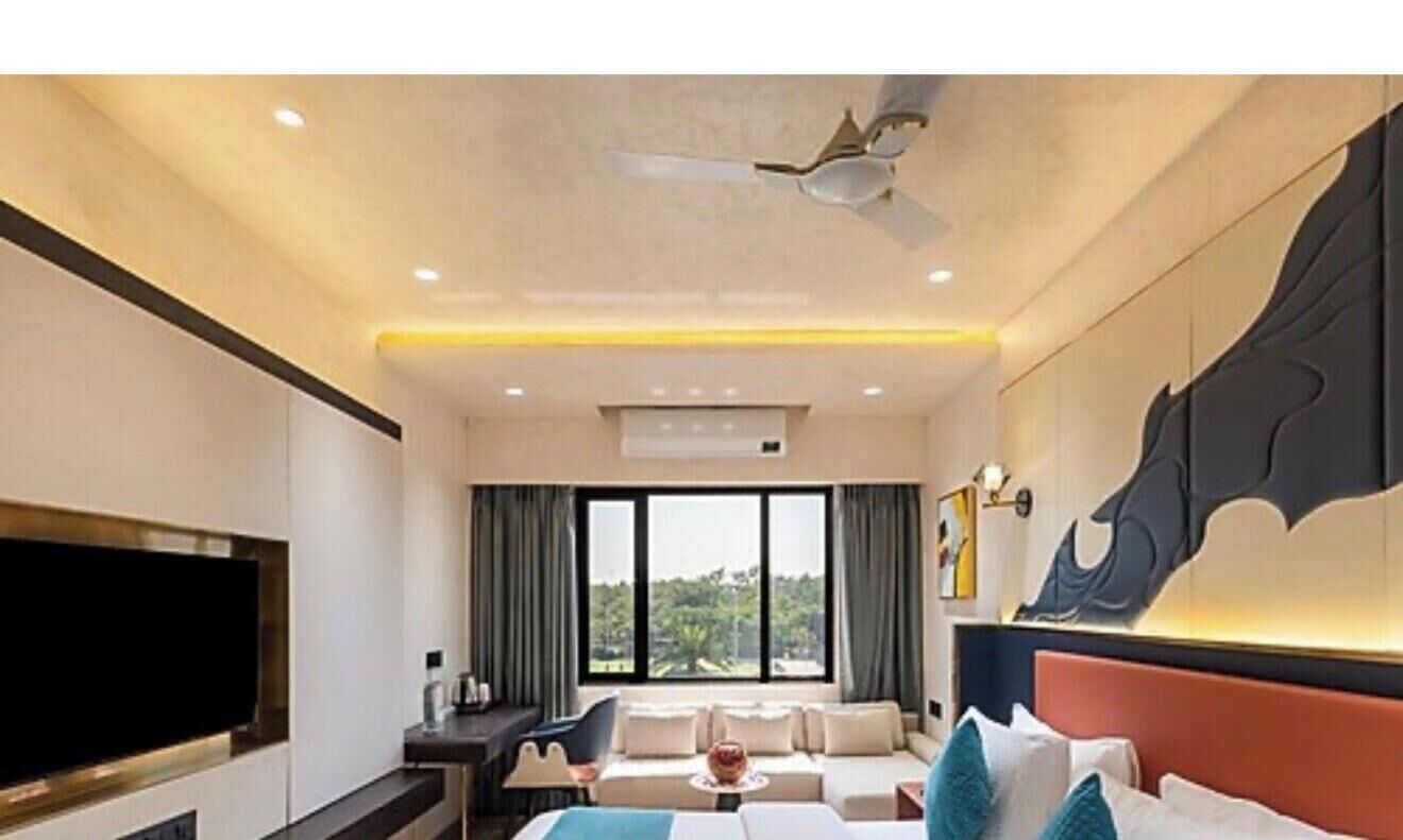 A hotel room with a large flat-screen TV, a bed with multiple pillows, a sofa, a desk with a chair, and a ceiling fan.