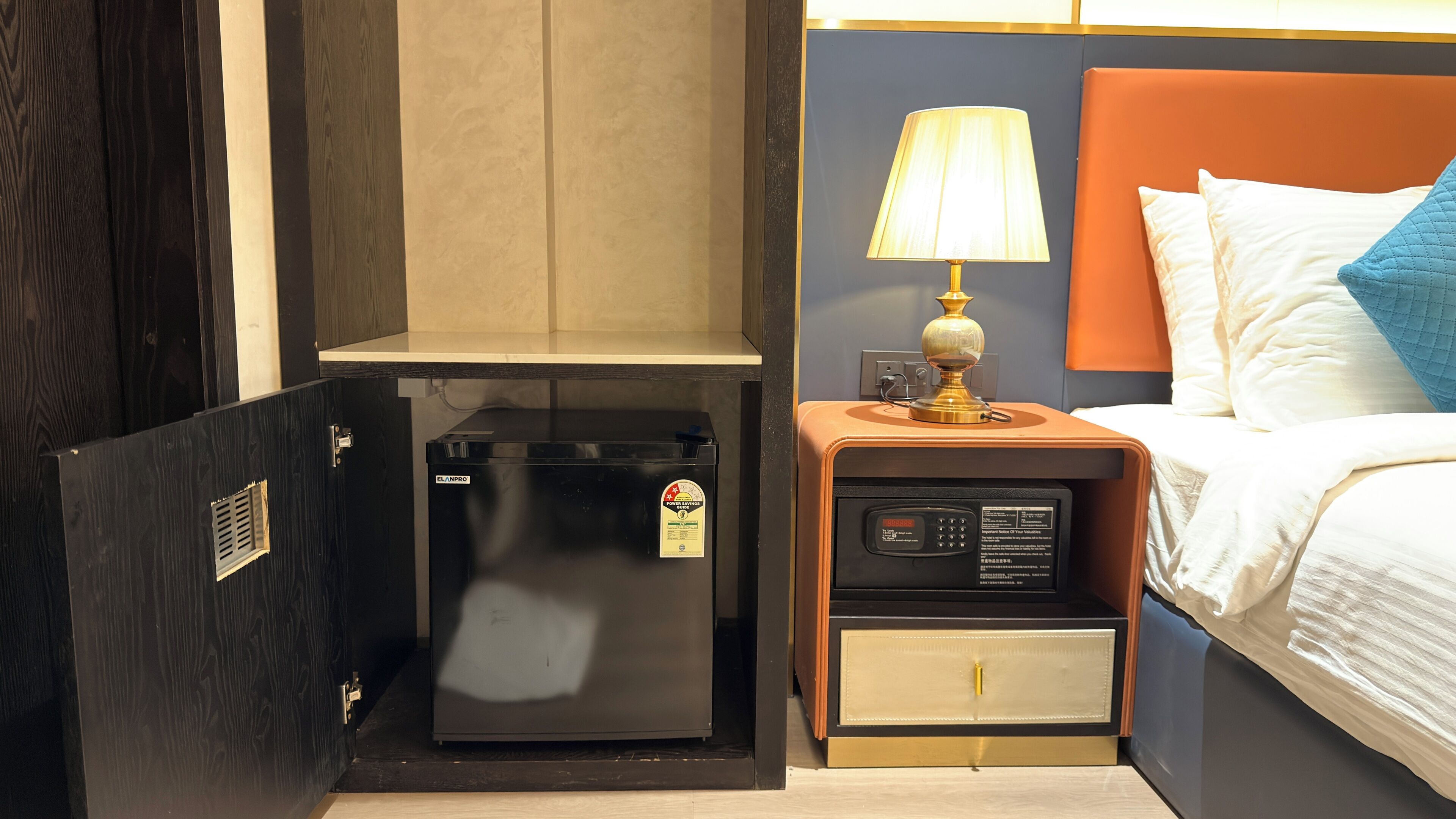 A compact room with a bed, a nightstand with a lamp, a small refrigerator, and a wardrobe.
