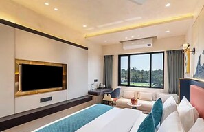 Habitación de hotel con una cama grande, un televisor de pantalla plana, un escritorio con una silla, un sofá y una ventana con vistas a zonas verdes.
