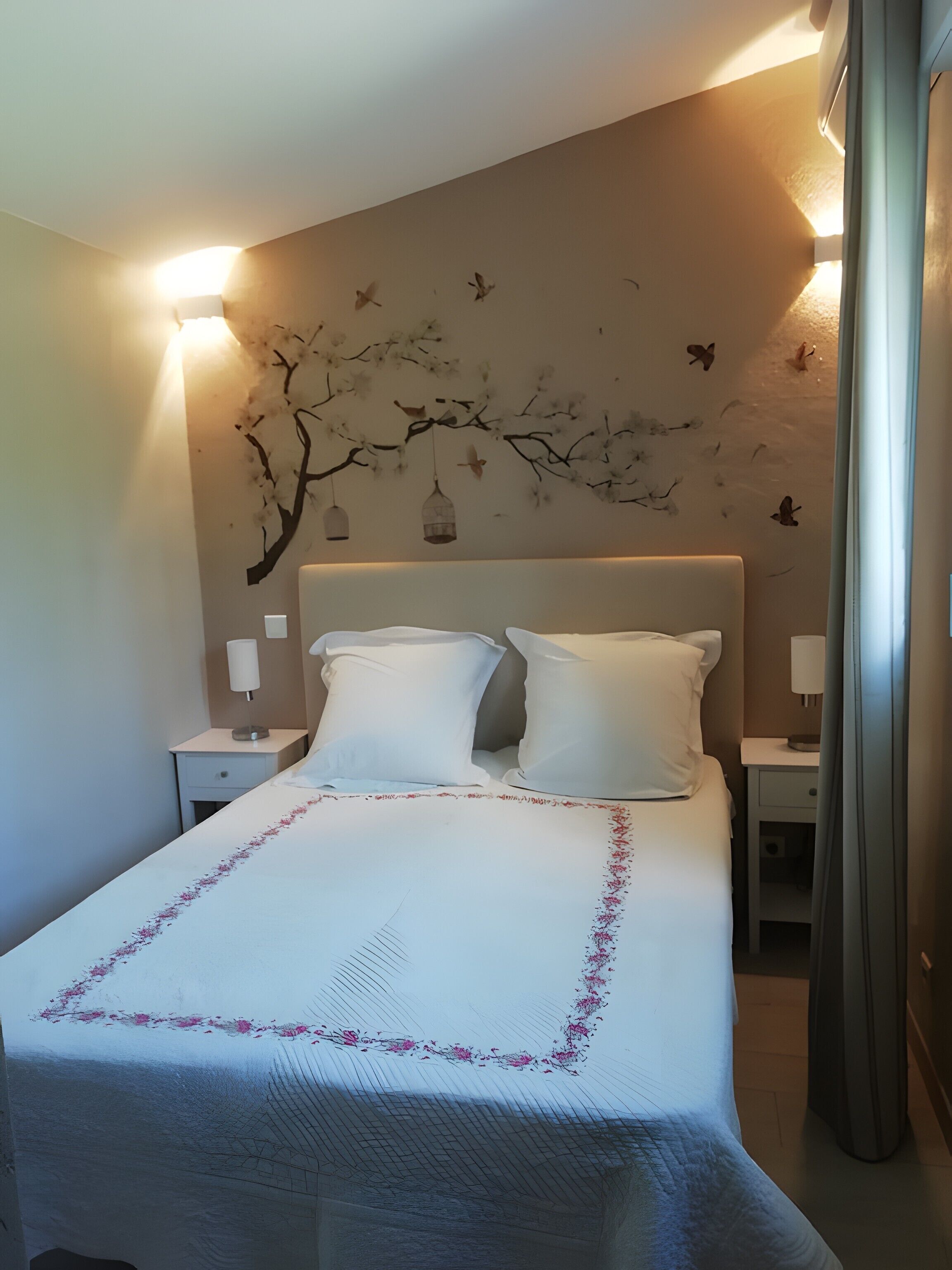 Een net opgemaakt bed met witte linnens en een roze bloemenrand, een muur met een muurschildering van een tak, en twee bedlampjes.