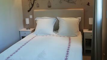 Een net opgemaakt bed met witte linnens en een roze bloemenrand, een muur met een muurschildering van een tak, en twee bedlampjes.