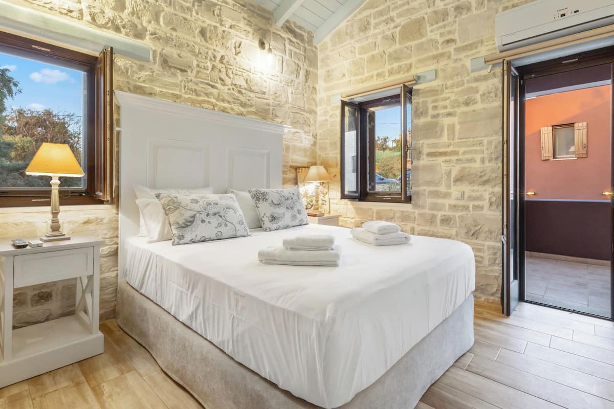 A bedroom with a stone wall, a bed with white linens, a nightstand with a lamp, and a view through a window.