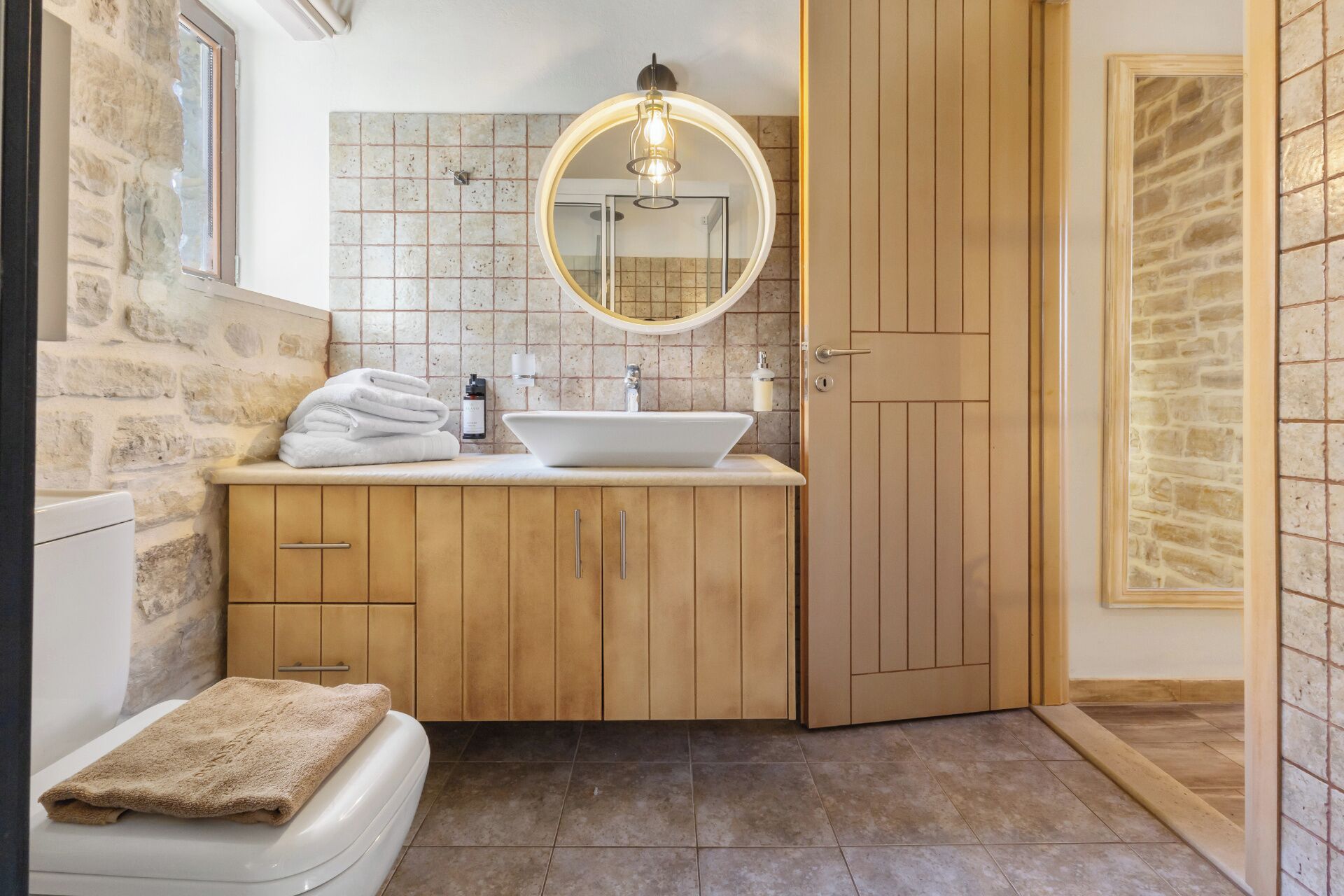 Une salle de bain avec un mur en pierre, un meuble-lavabo en bois, un lavabo blanc, un miroir et un porte-serviettes.