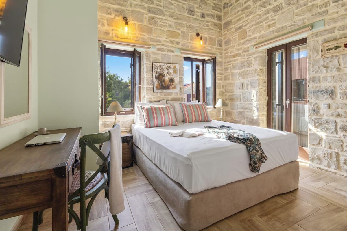 A bedroom with a stone wall, a bed with striped pillows, a desk with a laptop, and a window with a view.
