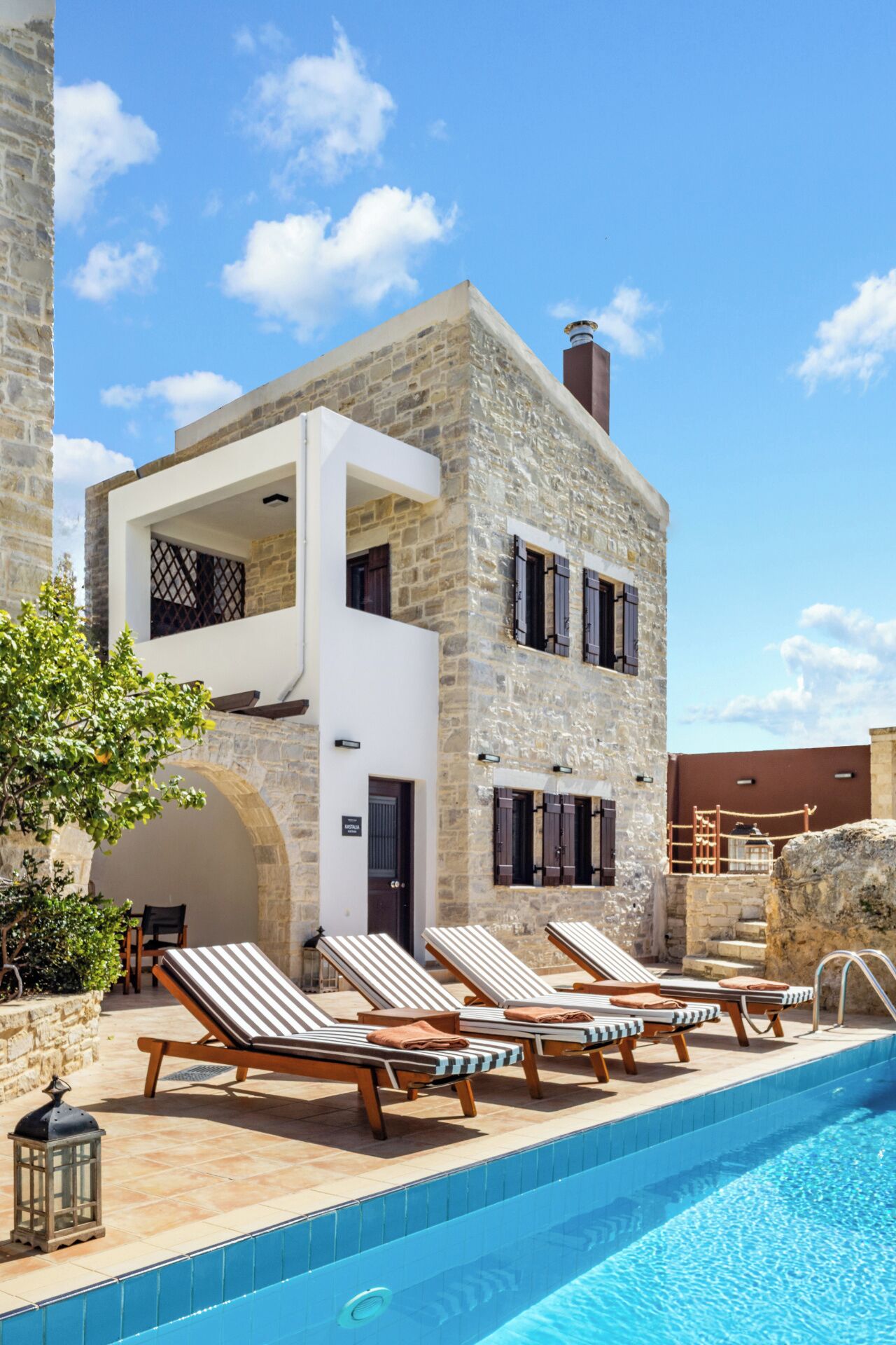 A two-story stone building with a pool, lounge chairs, and a clear blue sky.