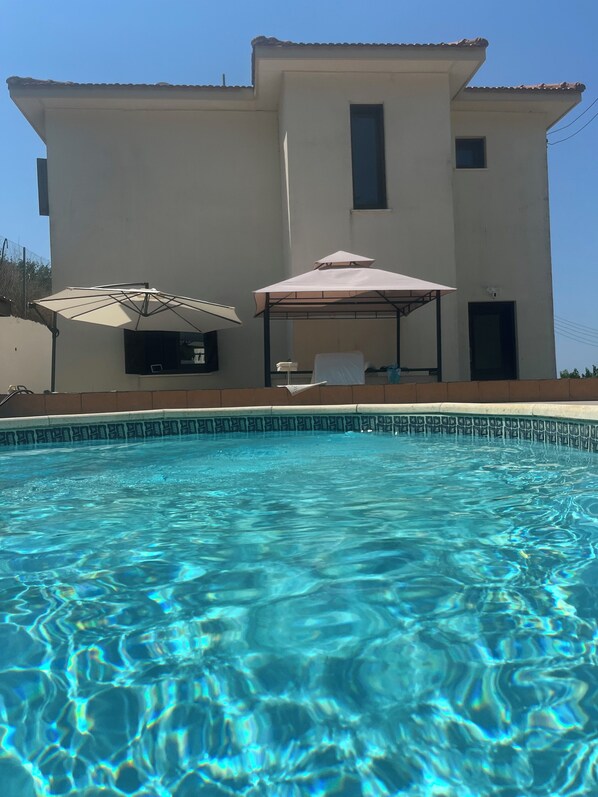 A swimming pool with clear blue water, a covered seating area, and a two-story building with a tiled roof.