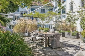 Une terrasse extérieure avec une table et des chaises, entourée d’une végétation luxuriante et offrant une vue sur un immeuble à plusieurs étages.