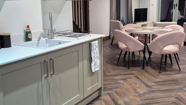 A modern kitchen with a central island, a dining area with pink chairs, and a herringbone wood floor.