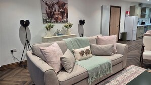 A living room with a sofa, two side tables, a coffee table, a painting, and a kitchen area in the background.