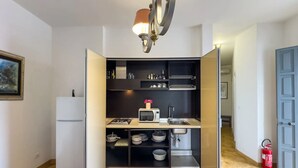 A compact kitchen with a gas stove, microwave, and a sink. There are shelves above the counter holding kitchenware and a small potted plant. A white refrigerator is to the left, and a fire extinguisher is visible on the right. - Villa Solaria - Giardino Privato and Aria Condizionata (Porto Cesareo)