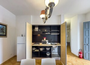 A compact kitchen with a gas stove, microwave, and a sink. There are shelves above the counter holding kitchenware and a small potted plant. A white refrigerator is to the left, and a fire extinguisher is visible on the right. - Villa Solaria - Giardino Privato and Aria Condizionata (Porto Cesareo)