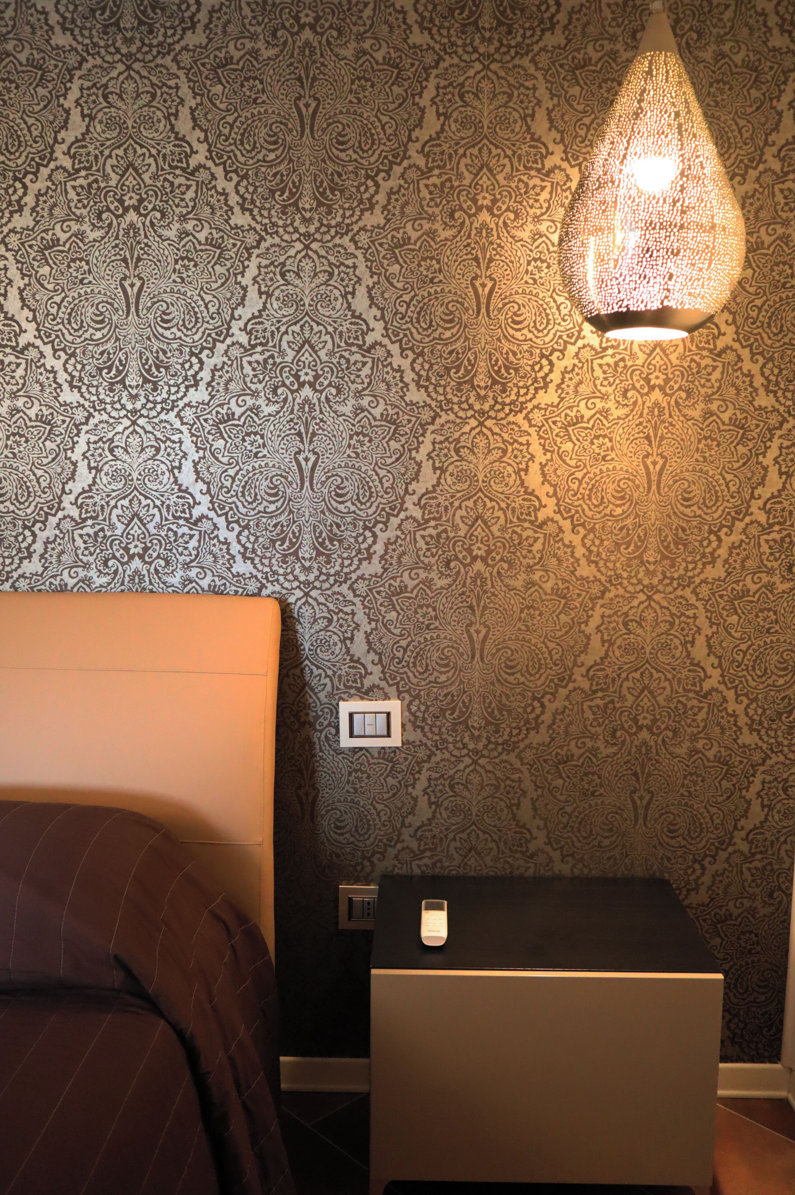 A bedroom with a patterned wallpaper, a bedside lamp, a nightstand, and a bed with a striped cover.