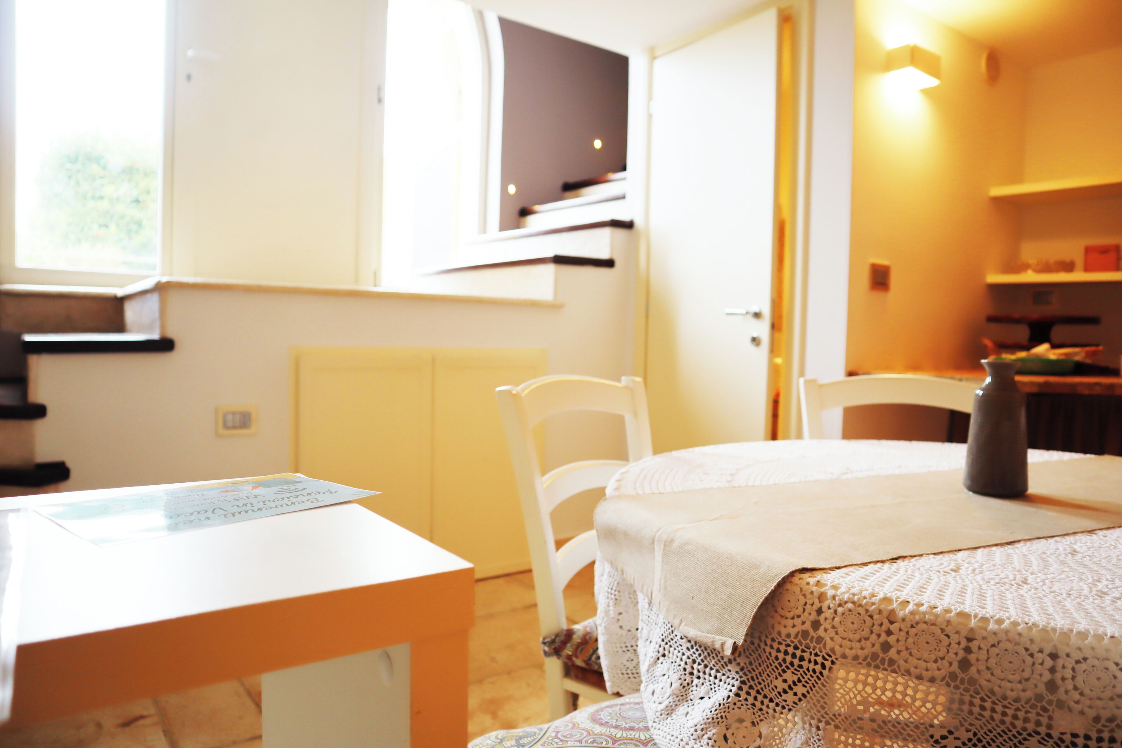 A room with a white table, a chair, a bed with a lace bedspread, and a window with blinds.