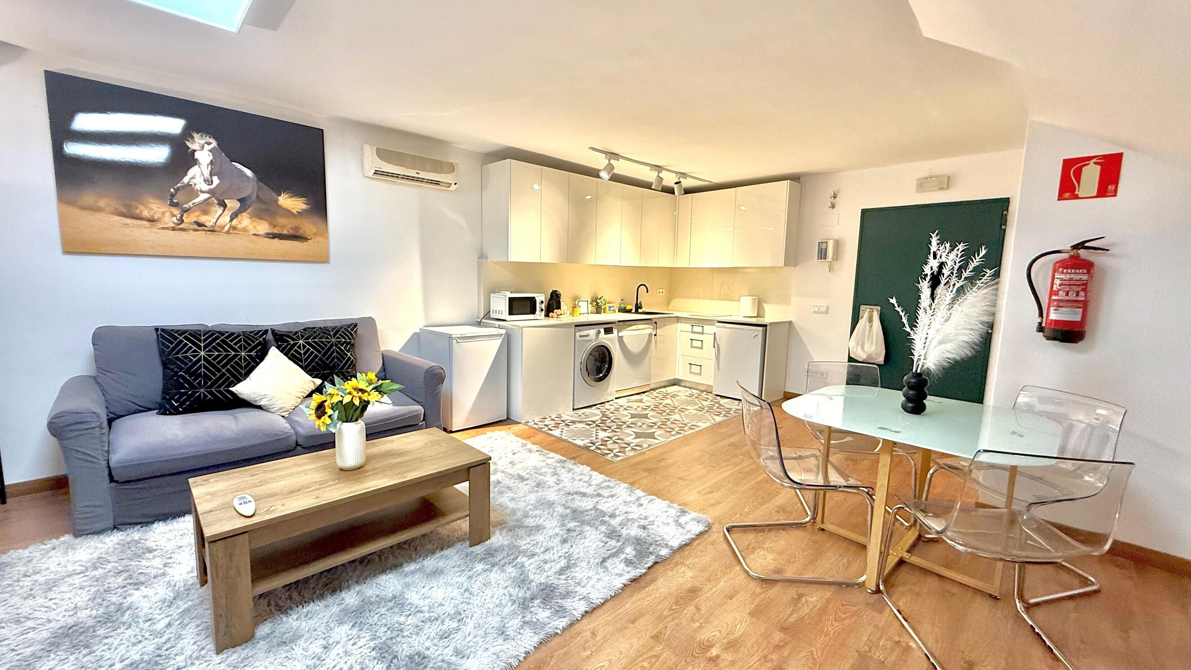 A compact living space with a grey sofa, a wooden coffee table with a vase of sunflowers, a glass dining table with chairs, a washing machine, a microwave, and a fire extinguisher on the wall.