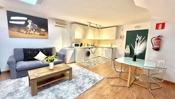 Een kleine leefruimte met een grijze bank, een houten salontafel met een vaas zonnebloemen, een glazen eettafel met stoelen, een wasmachine, een magnetron en een brandblusser aan de muur.