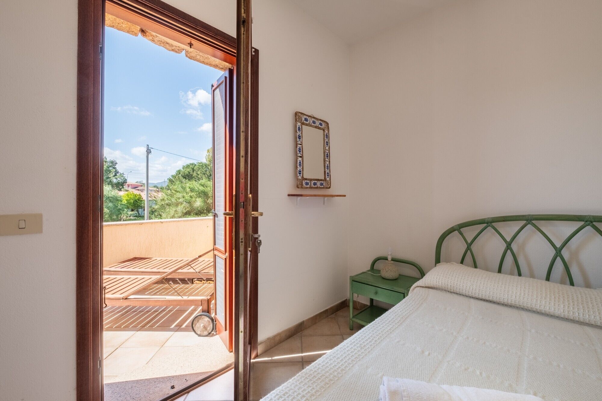Ein Schlafzimmer mit einem Bett, einem grünen Metall-Kopfstück, einem kleinen Nachttisch, einem an der Wand befestigten Spiegel und einer offenen Tür zu einem Balkon.