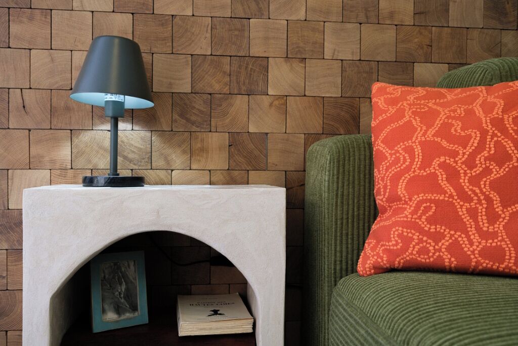 Rincón de una habitación con pared de madera, una mesita auxiliar blanca con lámpara, un sofá verde y un cojín estampado.