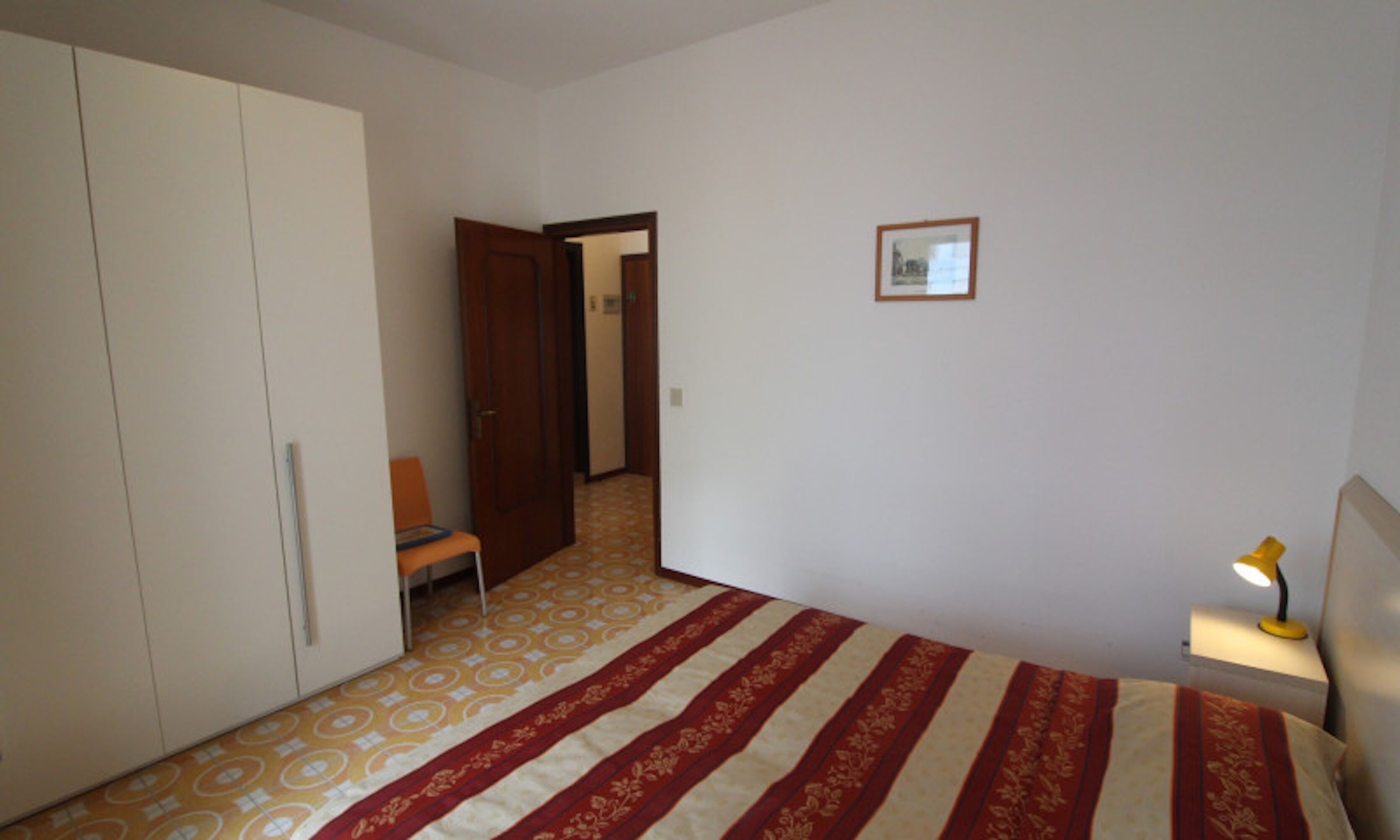 Un dormitorio con cama, armario, silla, mesita de noche con lámpara y puerta que da a otra habitación.