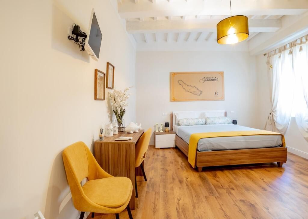 A bedroom with a wooden bed, a desk with a chair, a small table with a vase of flowers, and a map on the wall.
