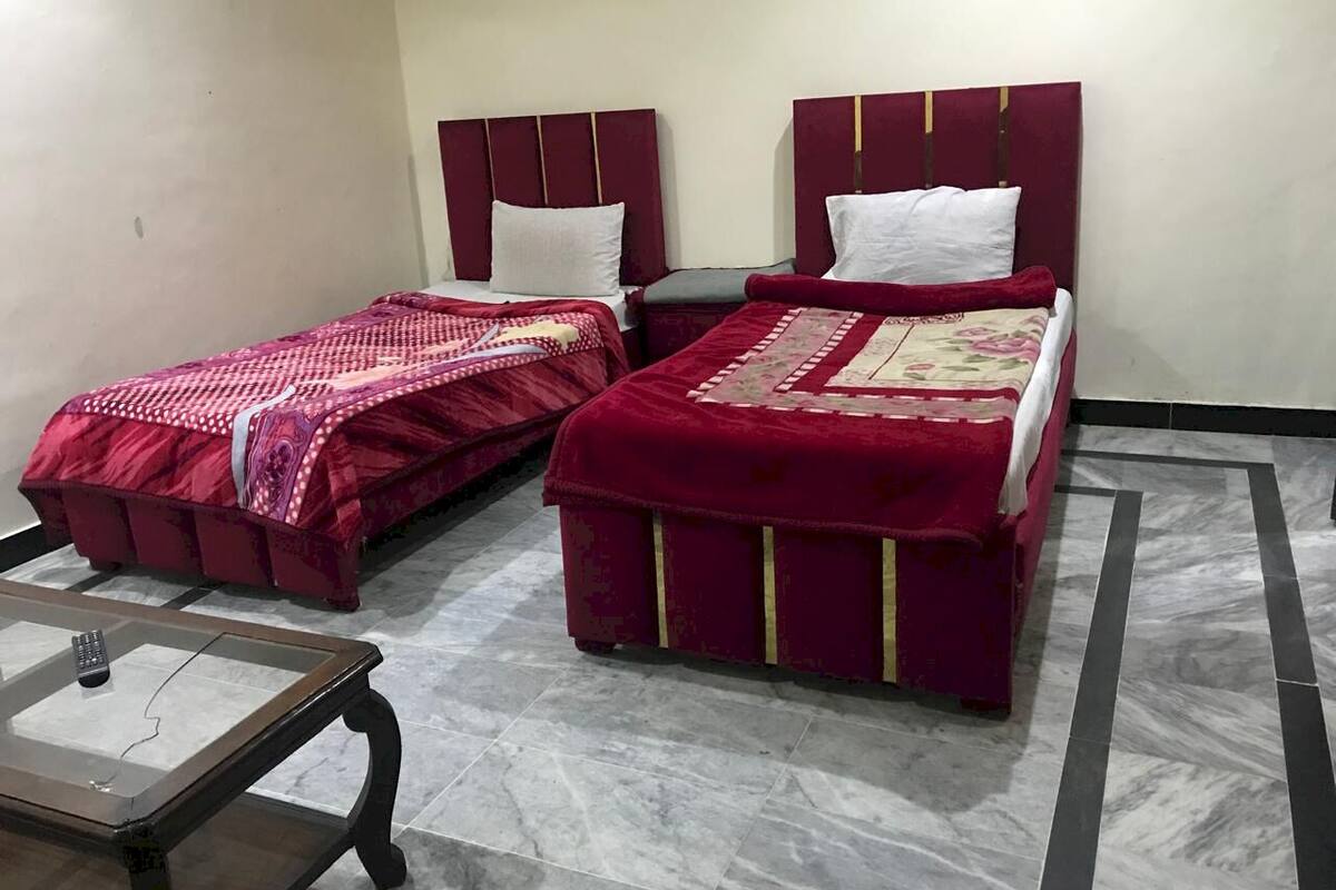 A room with two beds, each with a red bedspread and white pillows, a glass-top table with a remote control, and a patterned floor.