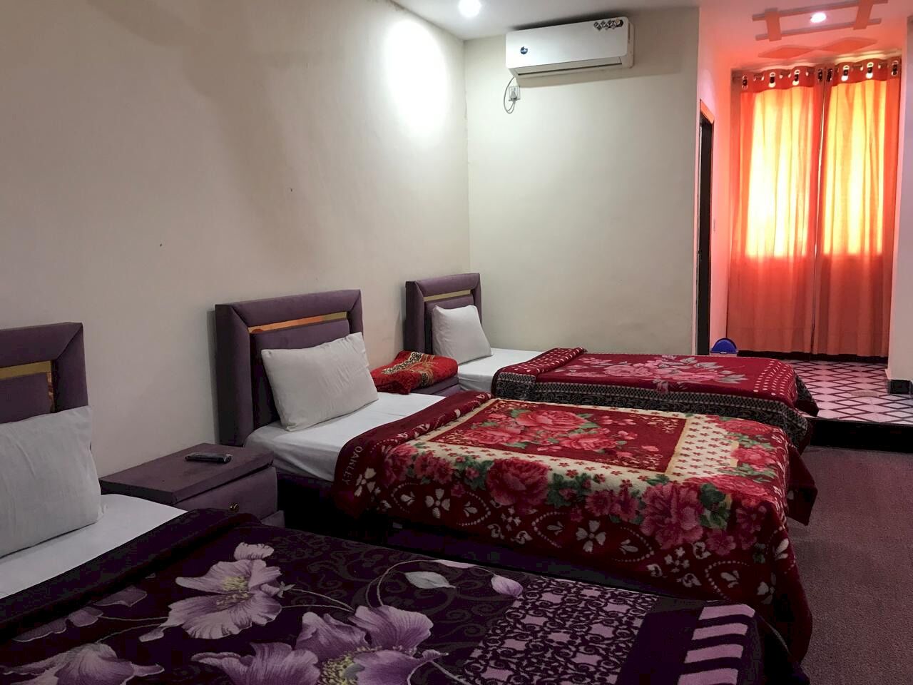 A hotel room with three beds, each with a distinct floral bedspread. There is a small table between the beds, and an air conditioning unit mounted on the wall.