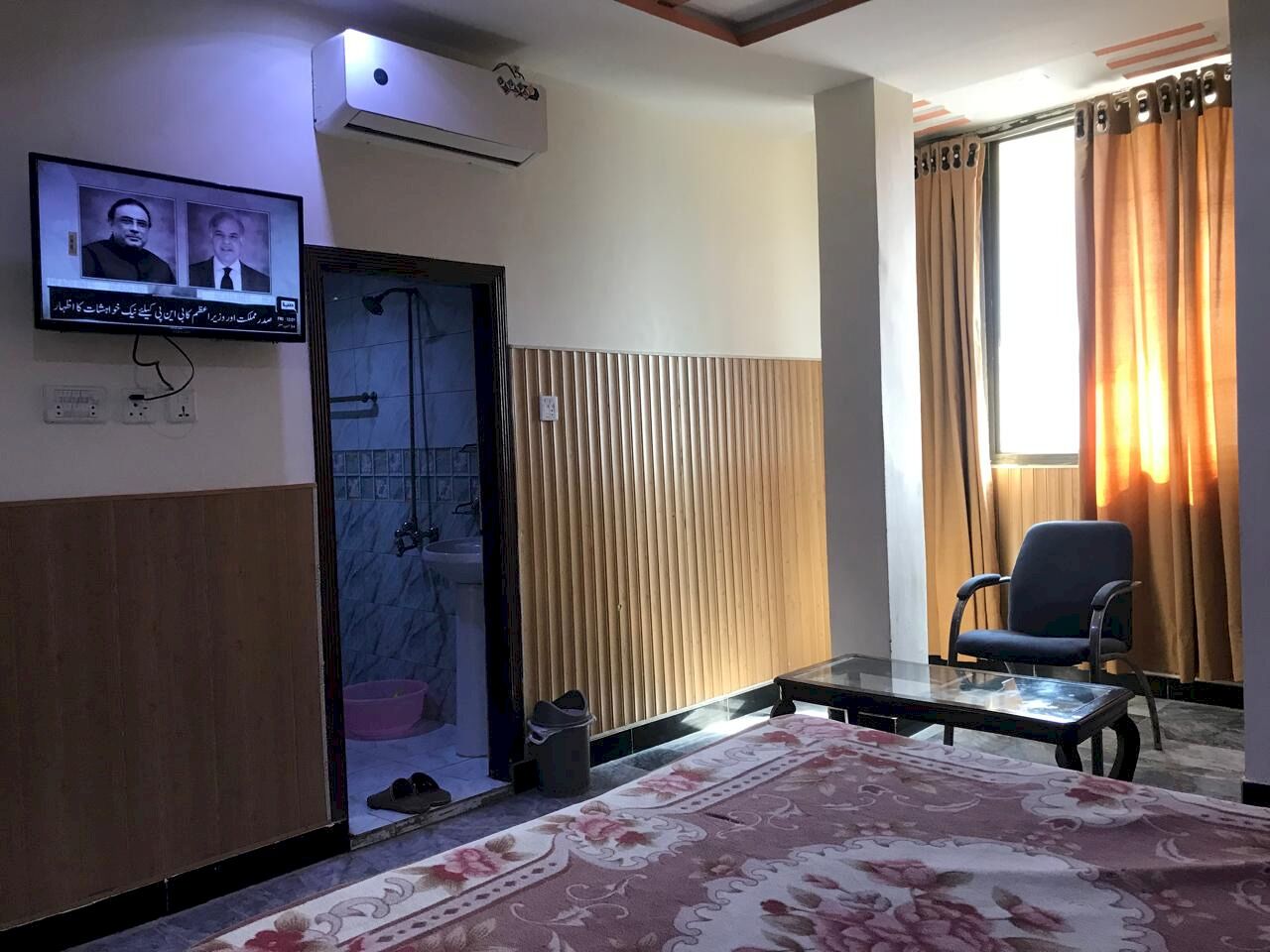 A hotel room with a bed, a television mounted on the wall, a window with curtains, a chair, and a small table.