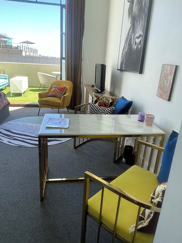 Et moderne hotelværelse med et spisebord, gule og blå stole, et lille fjernsyn og udsigt over bybilledet.