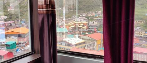 Ein Zimmer mit Bett, einem fenster mit Vorhängen und Blick auf eine Stadt mit Gebäuden und Bergen.