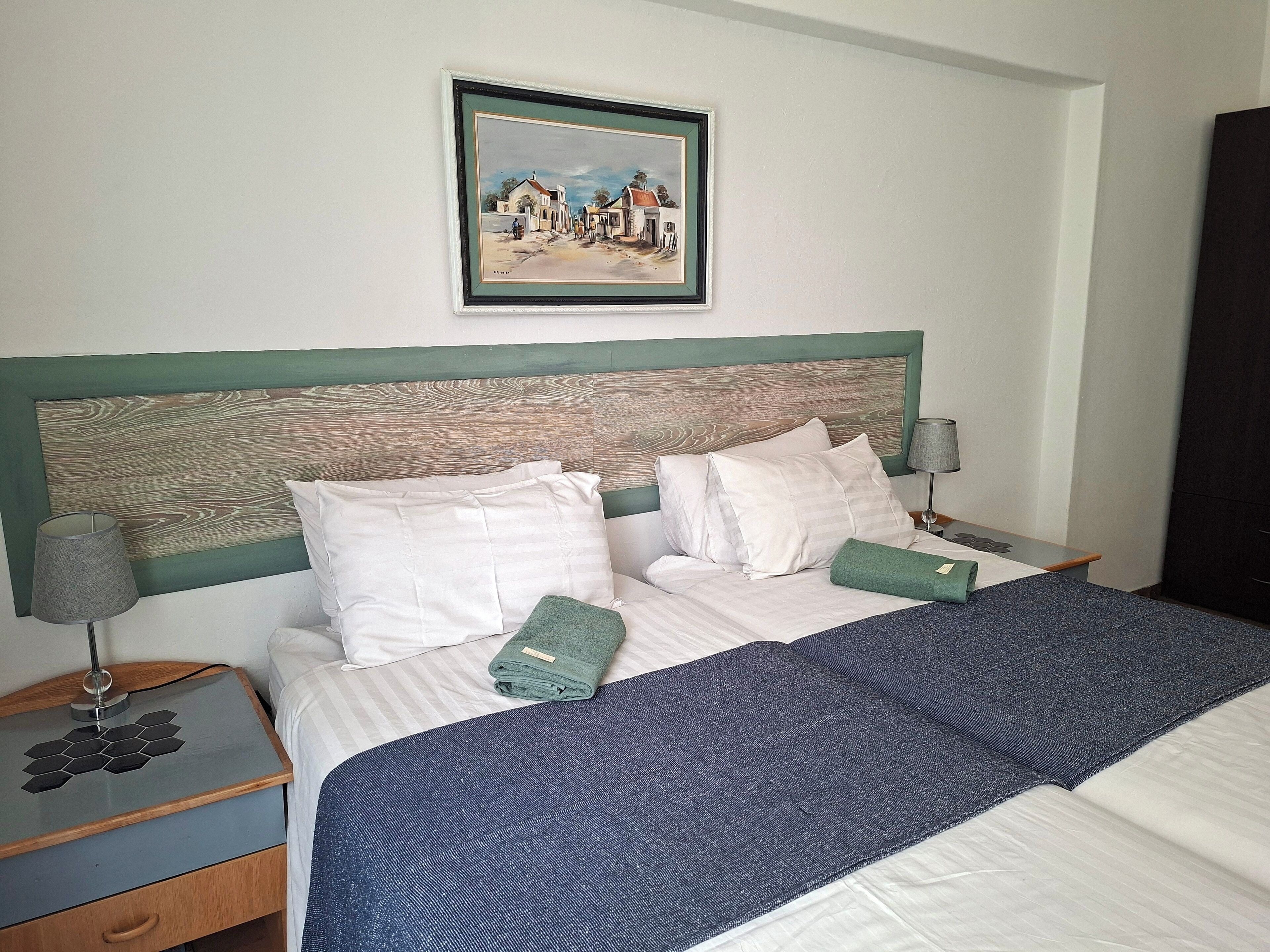 Ein ordentlich bezogenes Bett mit weißer Bettwäsche und einer blauen Decke, ein Holz-Kopfteil mit einem Bild darüber und ein Nachttisch mit einer Lampe und einer kleinen Schublade.
