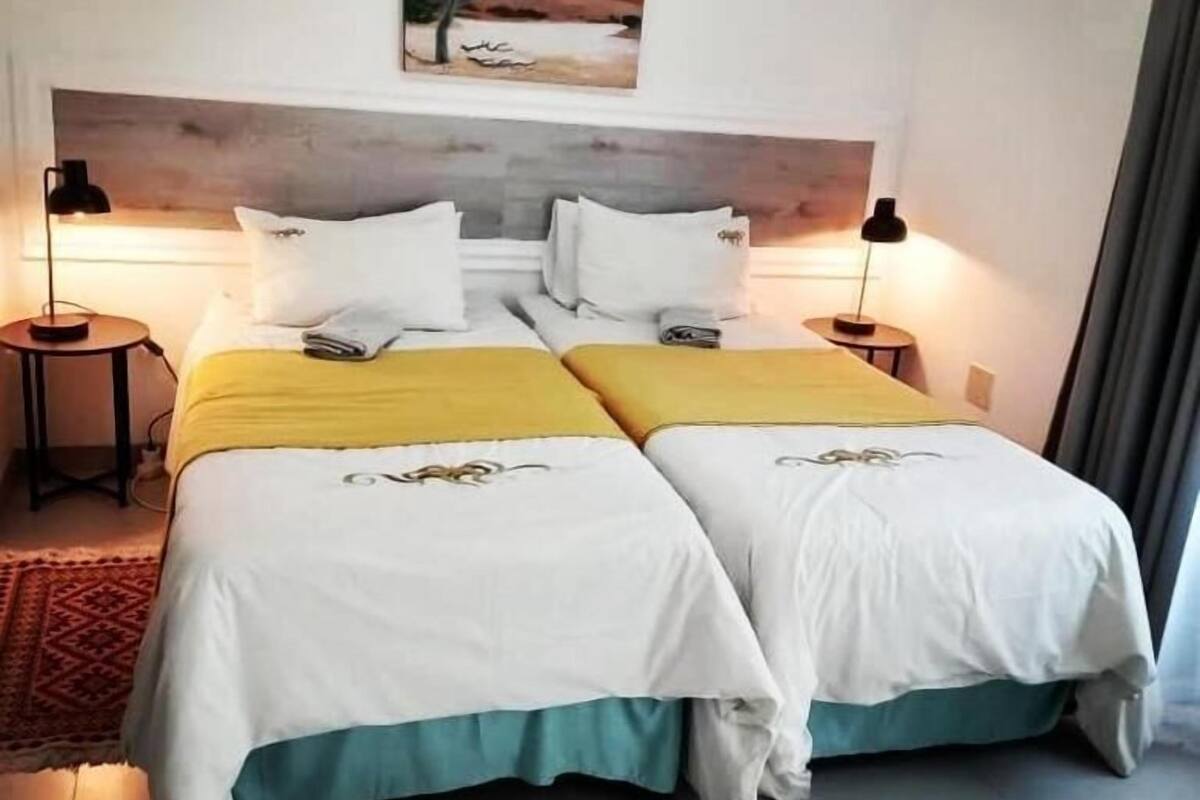 A double bed with white and yellow bedding, two bedside tables with lamps, a painting on the wall, and a view of the outdoors.