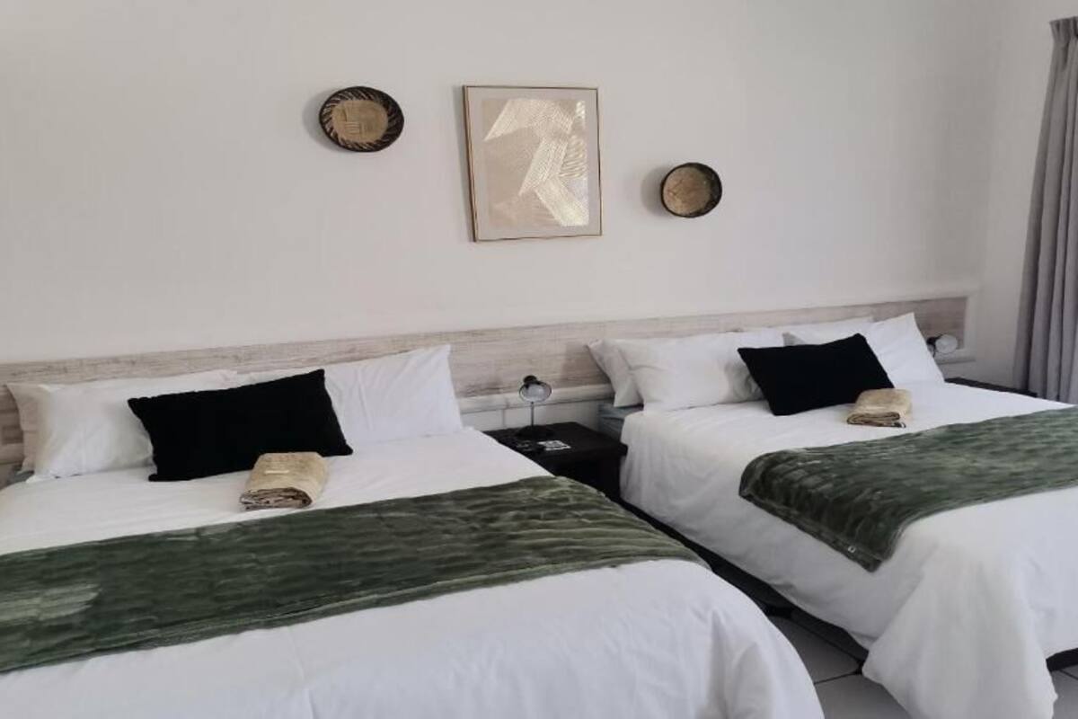 A hotel room with two beds, each with white bedding and a green throw blanket. There are two bedside tables with lamps, a wall clock, and a framed picture above the beds.