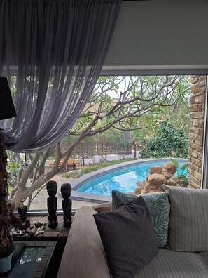 A room with a view of a swimming pool, a sofa with cushions, a lamp, and a sheer curtain.