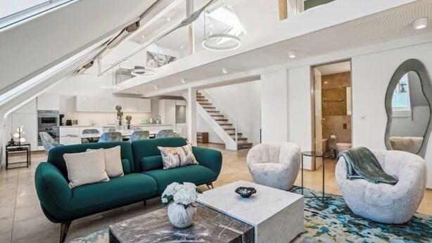 A spacious living area with a green sofa, a white armchair, and a marble coffee table. A staircase leads to an upper level with a kitchen and a bathroom visible. - Old Town Penthouse | 360° Views | Rooftop Terrace (Zürich)