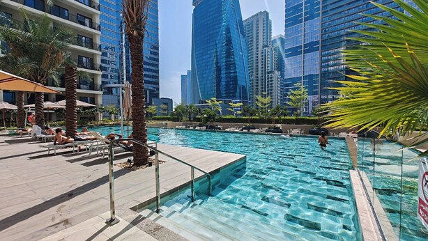 Ein Swimmingpool mit klarem Wasser, umgeben von Palmen und Liegestühlen, mit modernen Hochhäusern im Hintergrund.