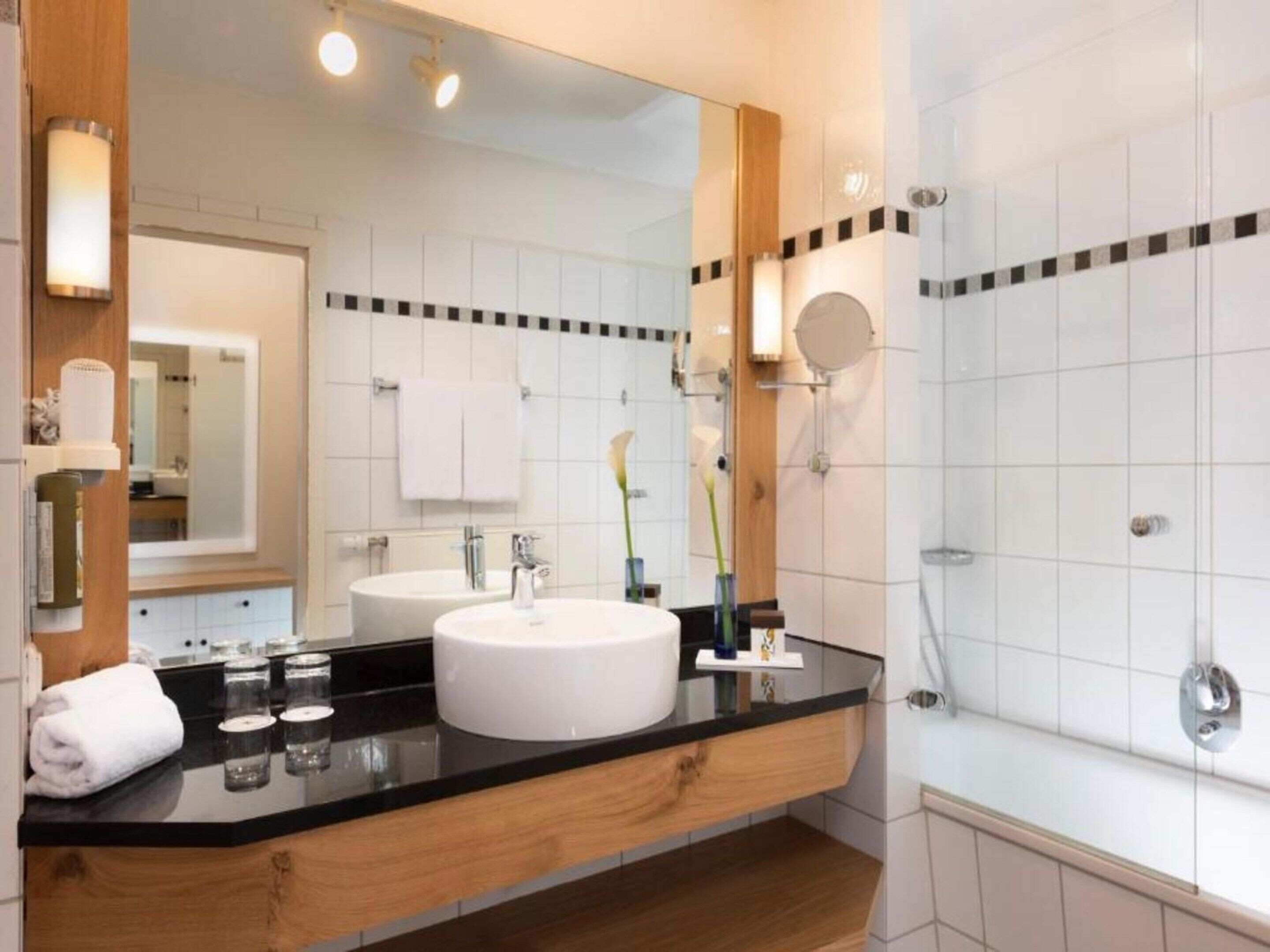 A modern bathroom with a large mirror, two sinks, a bathtub, and a vanity with a black countertop.
