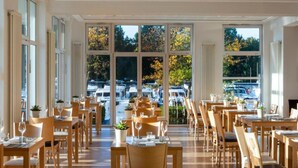 Un restaurante con mesas y sillas de madera, ventanas amplias y vistas a coches aparcados y árboles.