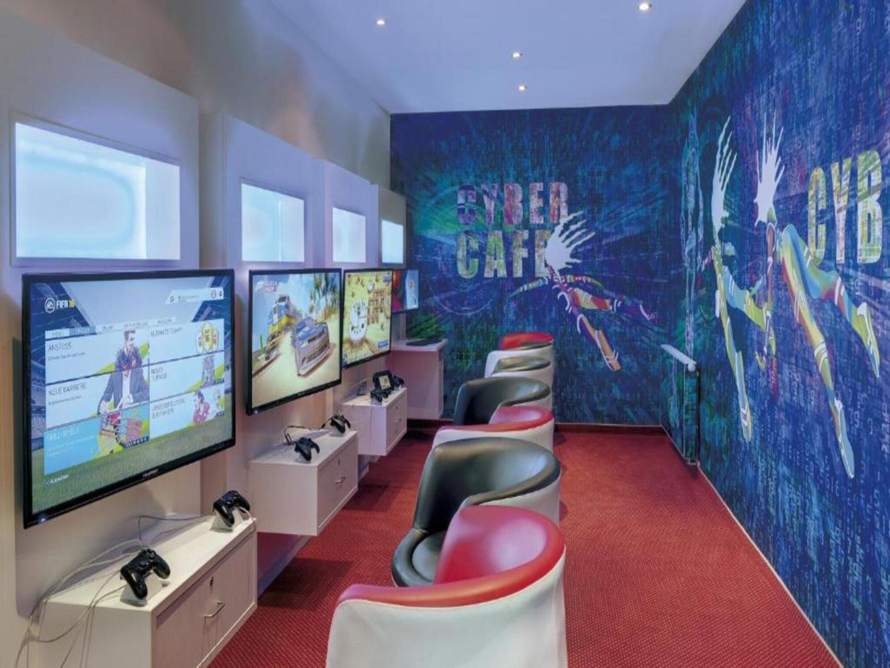 A gaming room with multiple screens, gaming chairs, and a mural with the text "CYBER CAFE".