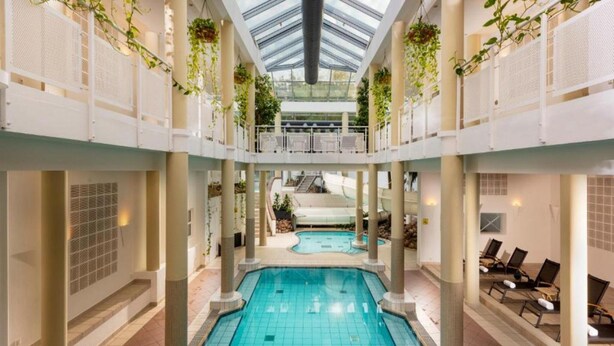 Una piscina interior con techo de cristal, rodeada de columnas y sillones de descanso.